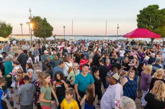 A crowd of hundreds of people dance on the rooftop of Monona Terrace with Lake Monona in the background