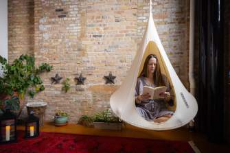 A white woman sits cross-legged and reads a book in a swinging cocoon chair inside Kosa Spa