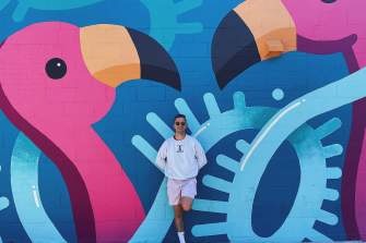 A white man stands with one leg bent against a large mural with blue lines criss crossing and large pink flamingos.
