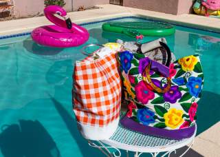 A colorful poolside scene featuring a vibrant, floral-patterned bag and a checked towel on a white metal table, with inflatable pool rings floating in the background.