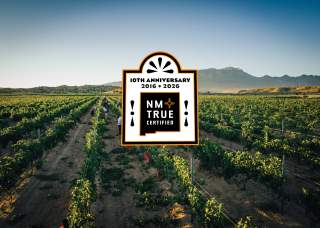 A vast vineyard stretches under a clear sky, with mountains in the background. In the center, there's a large "NM True Certified" logo celebrating its 10th anniversary from 2016 to 2026.