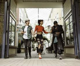 Three friends walk side by side, laughing in a bright, covered shopping walkway lined with storefronts.