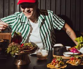 A person in a striped shirt uses chopsticks to pick up food from a table filled with tacos, greens, and drinks.