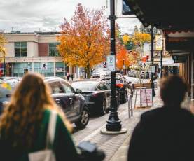 People walk along a downtown Nanaimo street with shops, parked cars, and vibrant autumn trees lining the road.