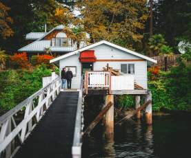 Two people stand on a narrow walkway leading to a small oceanfront house on stilts above the water, surrounded by trees and fall foliage.