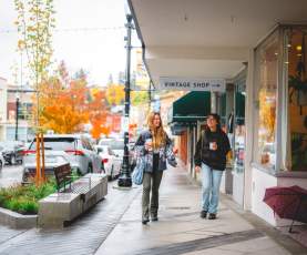 Two people stroll past shops on a rainy autumn day in downtown Nanaimo, holding coffee as colourful fall trees line the street.
