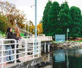 Two people pause along Nanaimo’s waterfront walkway, leaning on a railing and enjoying the view of the calm water and nearby park.