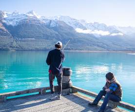 A man fishing off a wharf over a vibrant blue lake and snow covered mountains, while another sits and watches.