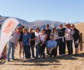 A group of people posing around a Love Queenstown sign with people tree planting and mountains in the background