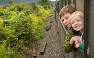 Two children leaning out of a train window with plant life to the left of the track.