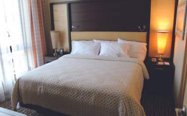 Hotel guest room with a king bed, neutral bedding, bedside tables with lamps, and natural light from a window.