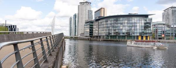 View of Salford Quays with modern glass-fronted buildings and high-rise apartments along the waterfront, a pedestrian walkway with railings in the foreground, and a small boat on the water under a partly cloudy sky.
