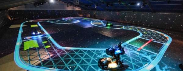 People driving electric go-karts on an indoor track with glowing neon lines and digital projections, creating a futuristic, space-themed racing environment.