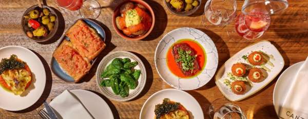 Overhead view of a wooden table filled with small sharing plates, including olives, grilled cauliflower, green peppers, croquettes and toast, alongside red drinks in glasses and a carafe, with “Canto” napkins and plates visible.