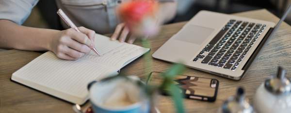 Person Writing On A Notebook Beside Macbook