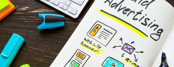 A keyboard and highlighters sit on a desk next to a notebook that has some drawings and the title, "Paid Advertising," on it.