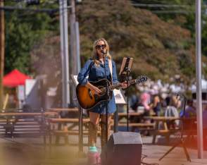 A female musician performing outdoors, playing an acoustic guitar and singing into a microphone at a local event with people and vendor booths in the background