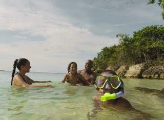 A family plays together in the water at a beach in Jamaica. In the foreground, a little boy with goggles and a snorkle looks at the camera.