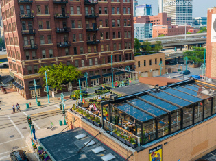 The Lux Skyhaus at Cafe Benelux, a glass greenhouse rooftop dining space in Milwaukee, overlooks a pedestrian plaza with teal lamp posts. The transparent structure reveals lush greenery and diners inside, surrounded by historic red brick buildings and downtown Milwaukee's skyline.