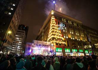 A lit Christmas tree display on the corner of a building and a stage below it with a crowd of people watching.