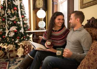 A man and a woman sitting on a couch looking at each other smiling as they're holding mugs in front of a decorated Christmas tree