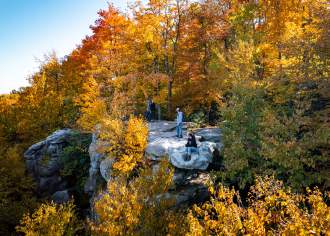 A girl sitting on the edge of a rock cliff with a man standing directly behind her with peak fall foliage surrounding them.