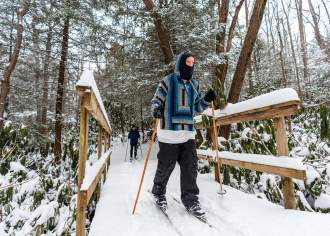 Two people in winter gear cross country skiing across a snow-covered, wooden bridge in the Laurel Highlands