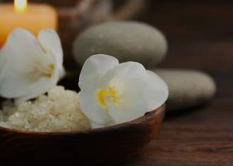 Spa essentials on a wooden surface featuring a bowl of bath salts, white flowers, essential oil bottles, stones, and candles for a relaxing retreat.