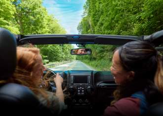 Two women in a corvette driving down an open road with green trees framing the road