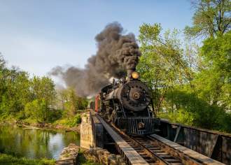 A large, historic train with exhaust coming out the top as it moves across train tracks above a river