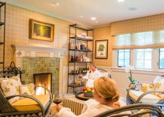 Two ladies in robes sitting in a luxurious sitting room at The Lodge at Glendorn with a lit fireplace. Both women are holding glasses of wine