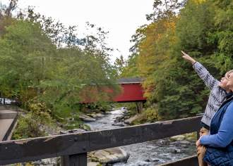 A woman carrying a young child as they stand on a bridge over a river with the child pointing up at something.