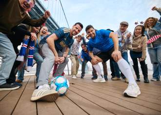 Two men in soccer jerseys squatting and posing for a photo with a soccer ball with a group of people cheering behind them