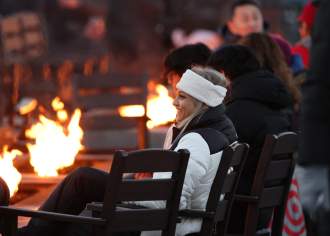 Woman in snow gear sitting around an outdoor firepit with other people.