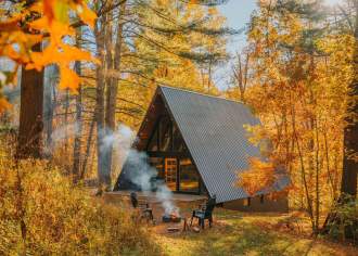 A cabin in a wooded area with yellow and orange trees and a fire pit in the front yard with four chairs around it