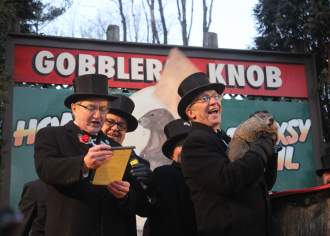 Three men in tops hats on a stage with a sign behind them that reads Gobblers Knob. One man is reading off a piece of paper while the other man stands behind him. The third man is smiling and holding a groundhog.