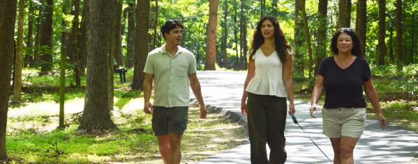 Three individuals walking along trail with trees in background, two have white shirts and one has black shirt.