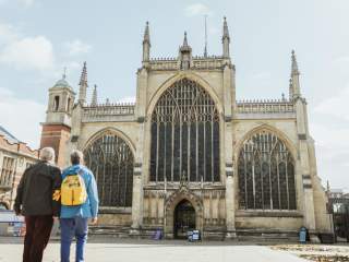 Two people stand outside a large Gothic-style church with tall arched windows, intricate stonework, and spires. The sky is bright and the church dominates the background.