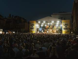 The stage at Live At The Yard at night. Razorlight are on stage infront of a banner emblazened with their logo. Bright lights are shining down on them and the crowd from the top of the pyramid shaped stage. Yellow banners flank both sides of the stage with Live At The Yard branding. In the foreground 100s of people are watching the band on stage.