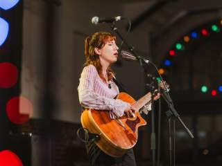 A woman plays an acoustic guitar onstage whilst singing into a microphone.