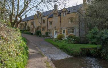 A row of traditional Cotswold stone cottages beside a narrow lane, with a shallow stream running alongside and trees and greenery in the foreground.