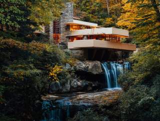 A modern, tan home , known as Frank Lloyd Wright's Fallingwater, situated on top of a waterfall with fall foliage-filled trees surrounding it