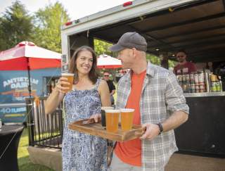 A man carrying a tray of beers and a woman walking with him with a beer in her hand at a festival