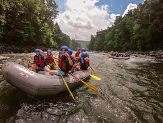 A group of people in helmets floating down a river in rafts. Two of the young boys are posing for the camera with a thumb up.