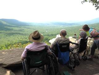 A group of people in wheelchairs sitting on an outdoor overlook looking at a scenic, green forest