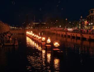 WaterFire Providence river night scene with a row of fire pits on concrete blocks, reflections in water, American flags, and spectators along the shore.