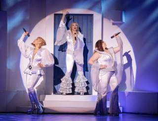 three women stand on stage with microphones in Mamma Mia