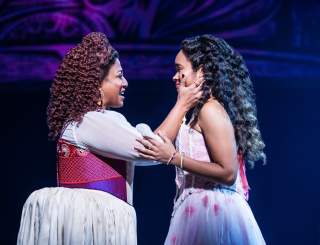 Two women stand on stage, the one on the left has her arm extended holding the face of the one on the right, smiling while she sings. The woman on the right holds the arms of the one on the left.
