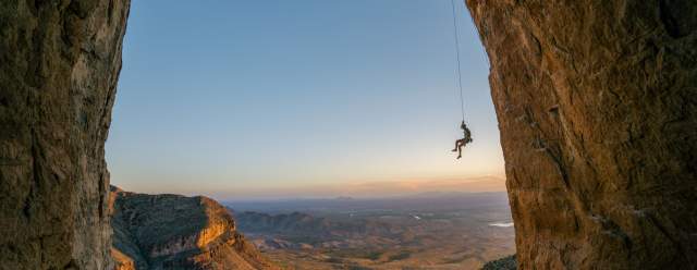 Climber Ben Hanna hangs from a rope anchored into a large arch, overlooking a horizon featuring cliffs and plains