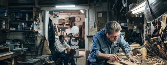 A person is working intently on a piece of wood in a cluttered workshop, while another person sits in the background holding the body of a guitar.